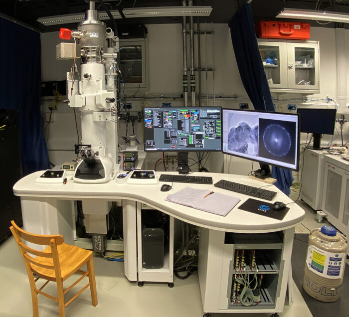 The new 100 keV electron microscope is set up, with computer monitors to its side and an old wooden chair in front, ready for use.