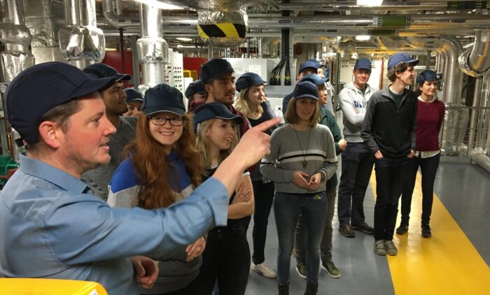 Stephen Holmes with a group of people wearing hard hats, surrounded by piping on one of the LMB’s interstitial floors