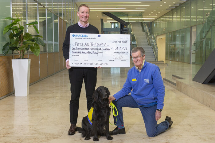 LMB Director Jan Löwe stands holding a large cheque with the fundraising total of £1414.95 on it. To his right, Charles, a Pets As Therapy volunteer, kneels, wearing a Pets As Therapy fleece and holding the collar of a black dog who is wearing a yellow volunteer jacket.