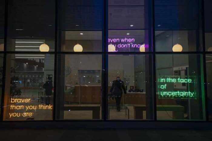 Display of three neon signs (one is orange on the left, the middle one is pink and the far right is in green) which display the messages of the art piece, Making Visible.