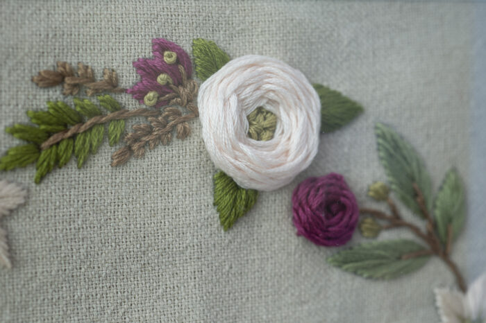 A close up of floral embroidery on a canvas fabric. Includes a large pale pink flower and a smaller dark pink flower wit green leaves.