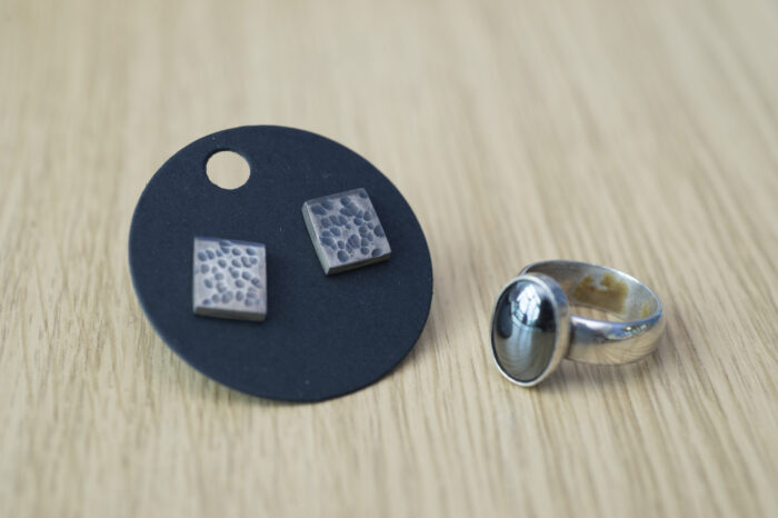 Two pieces of jewellery places on a wooden table. There are square earrings with  a speckled blue design on a round stand, and a sliver ring with a dark inset gemstone.