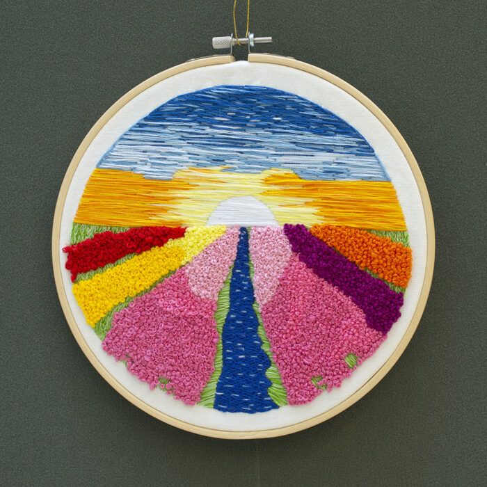 An embroidered landscape on a embroidery hoop. The landscape shows a blue and orange sky with a sun on the horizon. There are red, free, yellow, pink, blue, purple, and orange stretches of land receding to the horizon line.