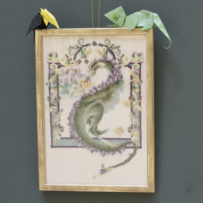 A framed rectangular cross stitch of a green dragon with flames coming out of its mouth. The dragon is framed within a stitched border of purple, yellow, blue and green design. An origami toucan  and chameleon are placed on top of the framed tapestry.