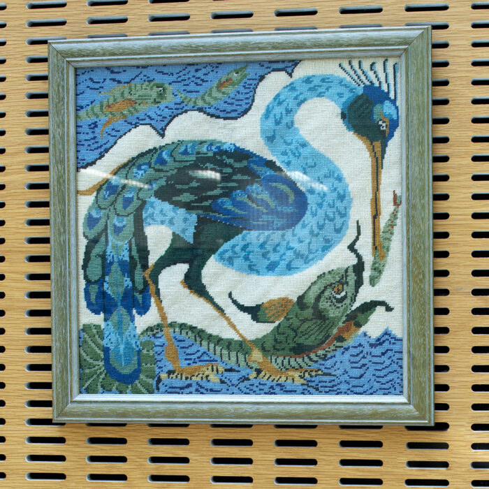 A framed square tapestry of a peacock and fish. The peacock is blue against a white backdrop. The fish are green and yellow against a blue backdrop.