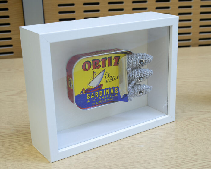 A white box frame displaying a Ortiz sardine can with crocheted sardines poking out of the pulled back lid.