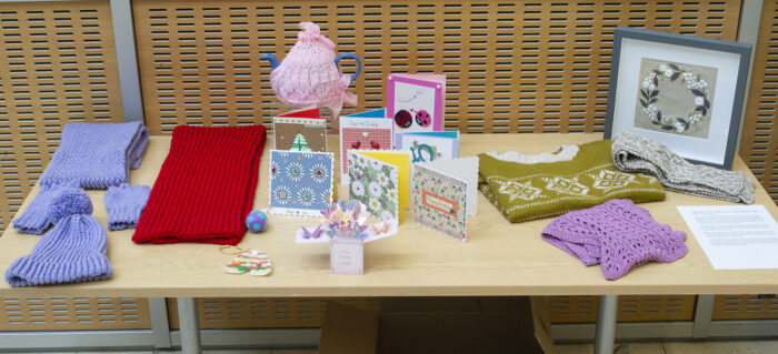 Various knitted items displayed on a table including a blue hat, scarf and gloves set, a red scarf, tea cosy, green and white fair isle jumper, lilac garment, and grey scarf. There are also hand-made cards displayed and a framed piece of embroidery.