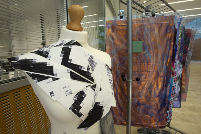 A mannequin stand with a black and white garment on display in the foreground. Behind it, textiles samples are visible on a railing. 