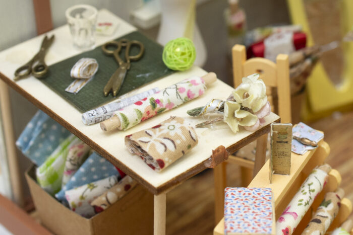 A close up of the interior of a miniature model room. There is a miniature table with fabrics, scissors, cups, and yarn displayed.