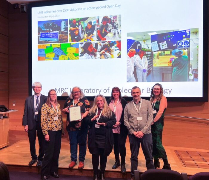 A group from LMB’s Biological Services Facility stands smiling on a stage, displaying a glass trophy and framed certificate. Behind them a large screen shows photos from LMB engagement activities.