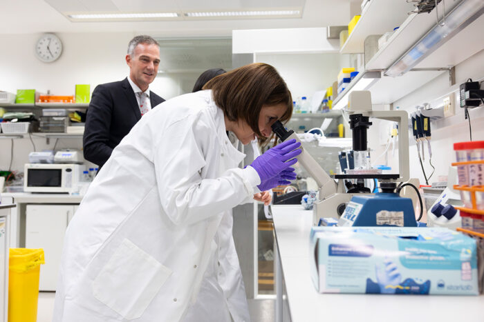 The Chancellor, Rachel Reeves, wearing a white lab coat and purple gloves, looks down a microscope at a lab bench. Behind her is stood Peter Kyle, Secretary of State for Science, Innovation and Technology.