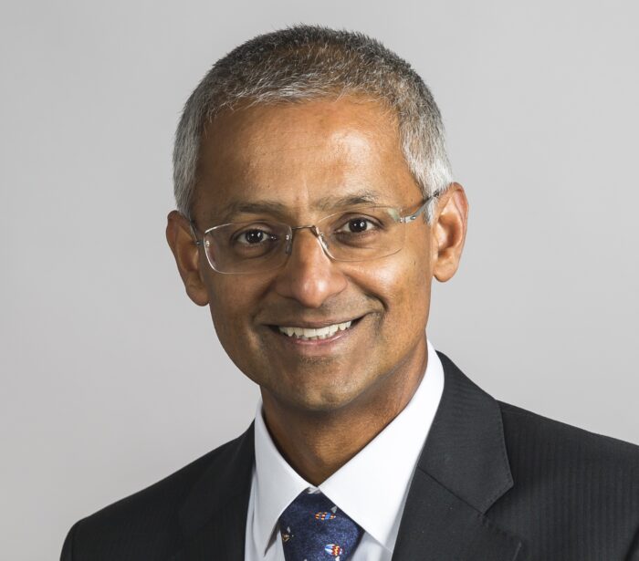 Shankar Balasubramanian, smiling wearing a suit and tie against a plain, grey background