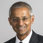 Shankar Balasubramanian, smiling wearing a suit and tie against a plain, grey background