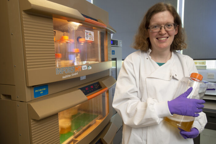 Keren Turton, wearing a white lab coat and purple gloves, holds a bottle of biological media